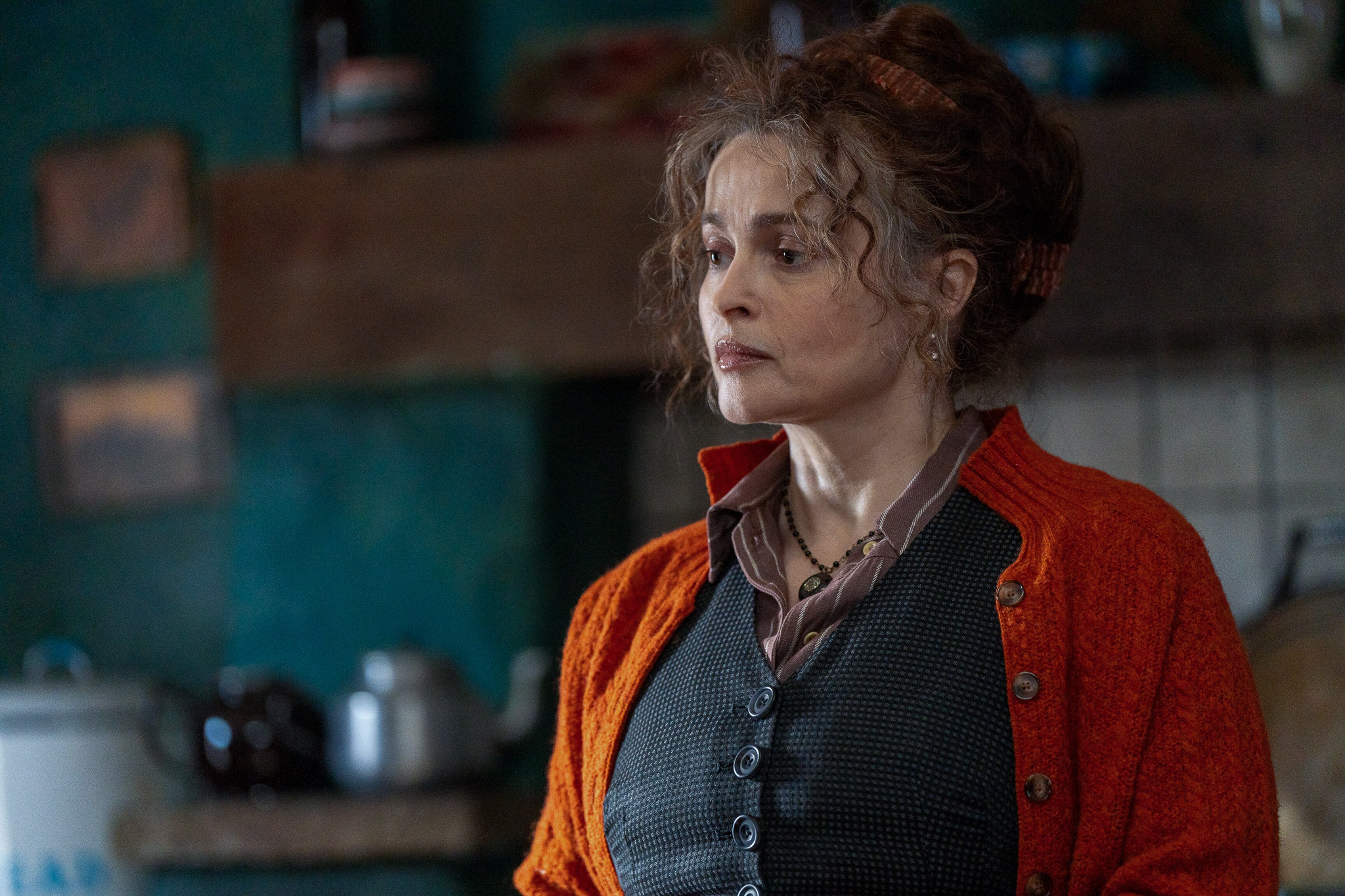 Helena looks to the left while standing in a darkened blue kitchen, wearing an orange cardigan and dark vest.