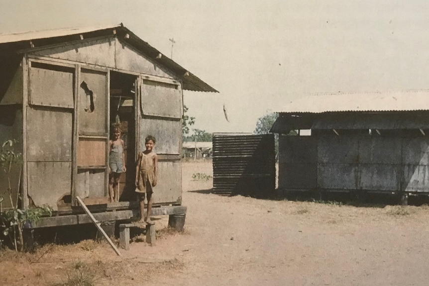 Two children stand in the doorway of a small timber building.