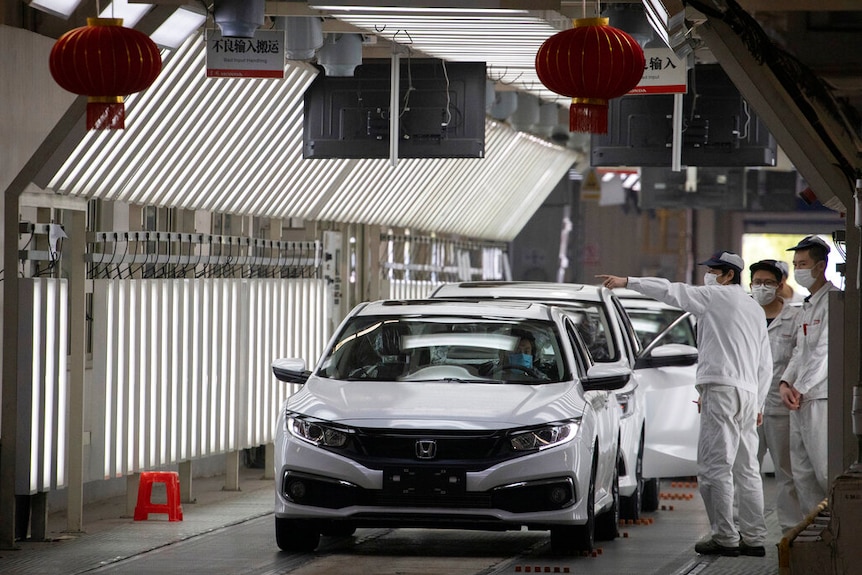 Workers wearing masks stand next to a production line of white Honda hatchbacks.