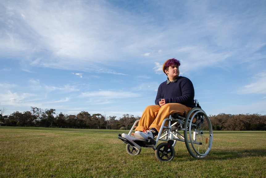 A young man in a wheelchair sits in the middle of a large, green-grassed oval.