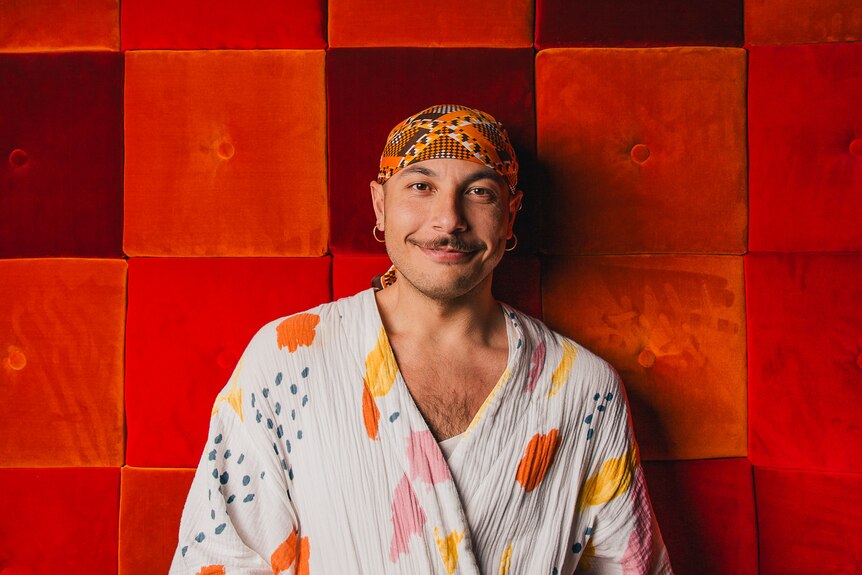 Non-binary person with brown moustache wears orange patterned bandana and cape with colourful splotches against orange wall.