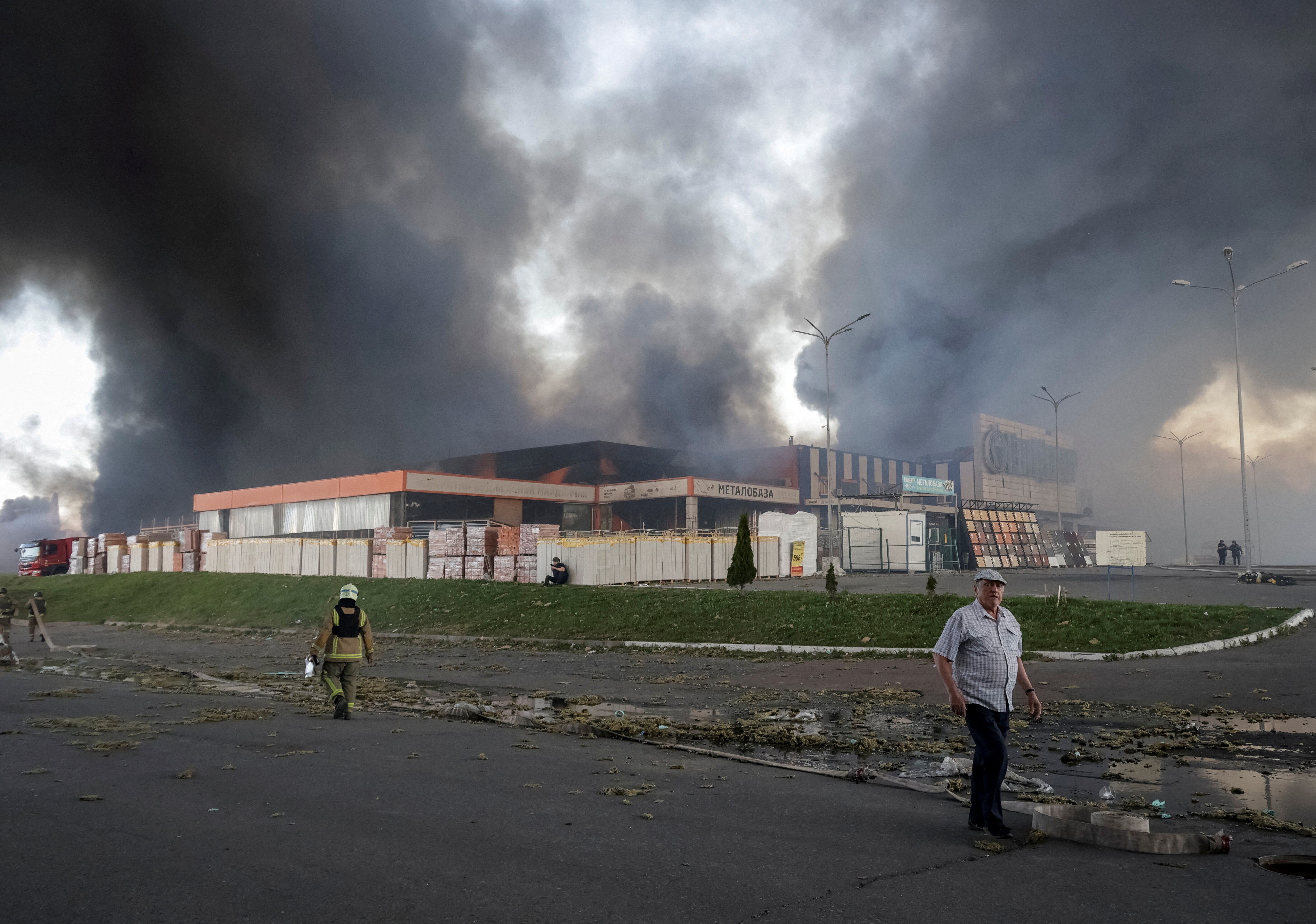 A firefighter and a man stand in front of a burning store, with smoke flowing into the sky.
