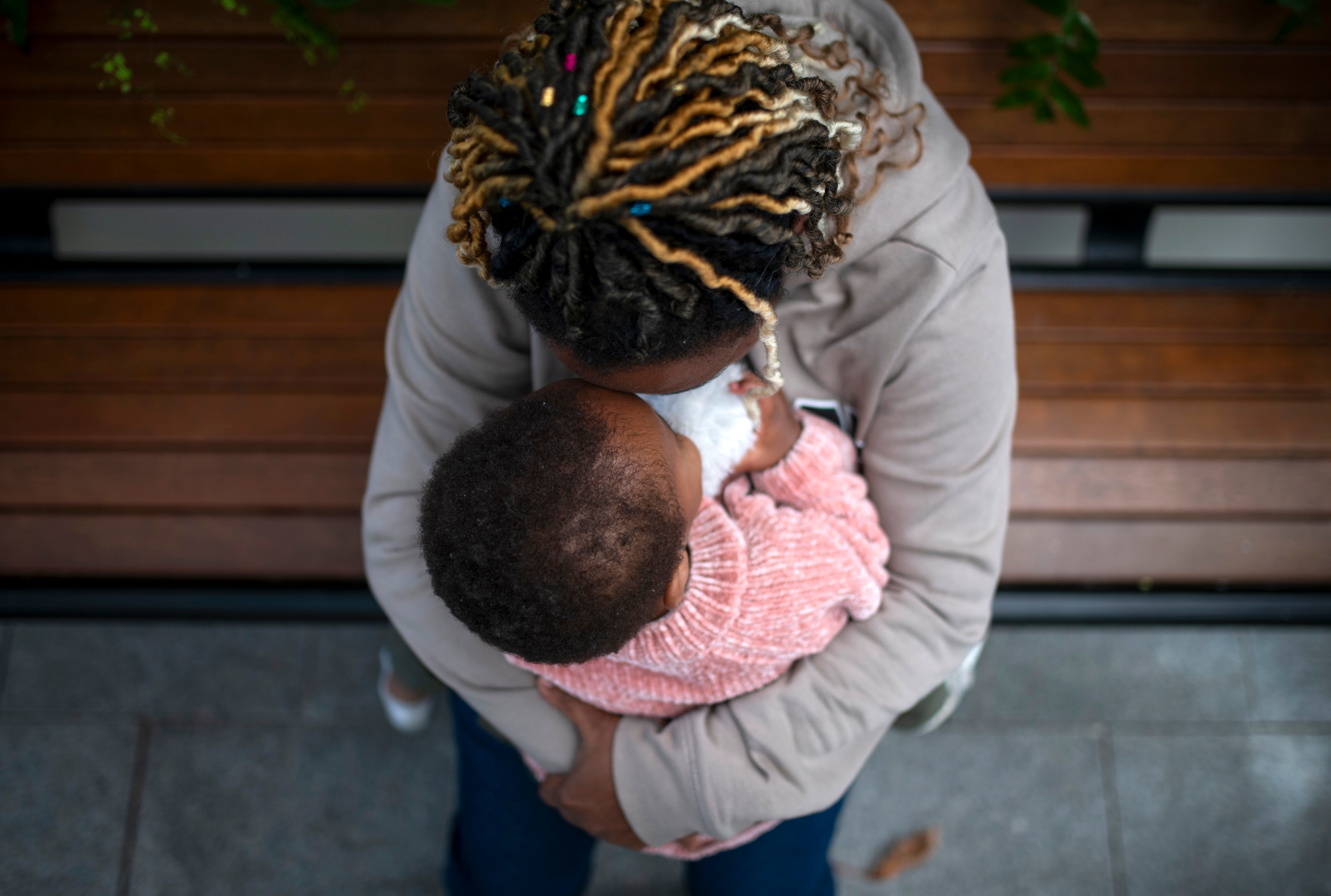 A woman in a grey hoodie holds a little girl in a pink hoodie on a wooden park bench from above.