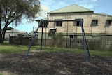 An unused swing set in a children's park, with an old house with flaking pain the background.