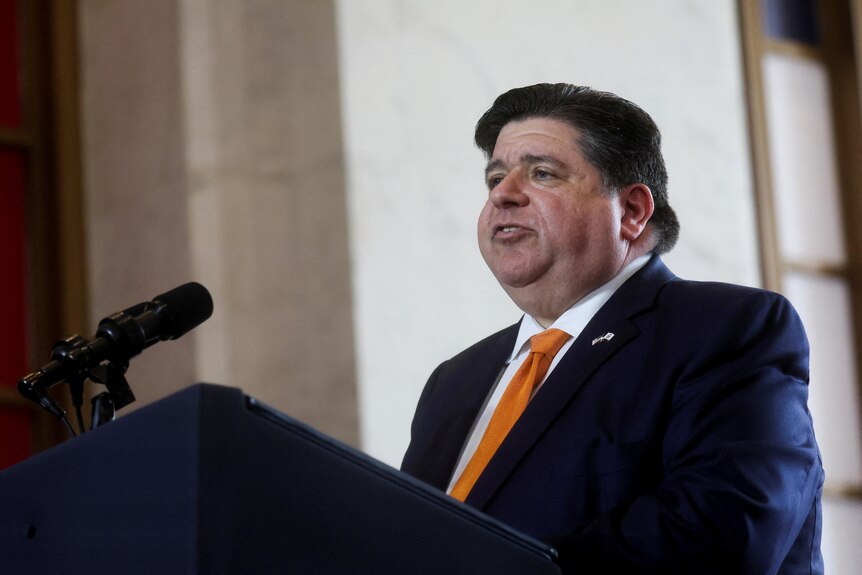 A man in a suit with an orange tie speaks at a podium 