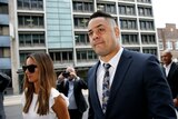 Jarryd Hayne walks in a dark suit with a floral tie next to a woman in sunglasses and a white dress.