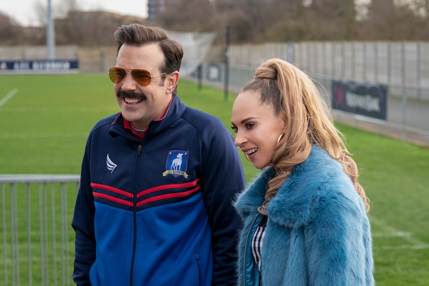 A moustachioed 40-something soccer coach and a 30-something woman with a high ponytail stand together on a soccer field
