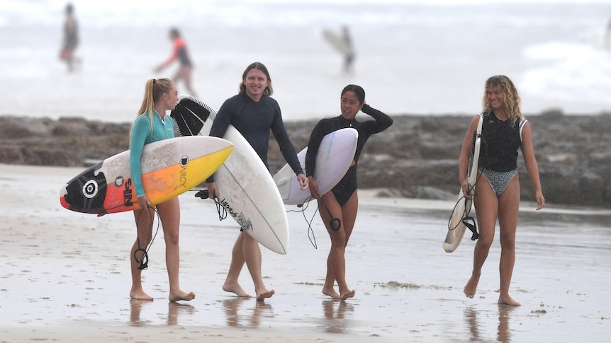 Four surfers in swimsuits and carrying surfboards walk along a beach
