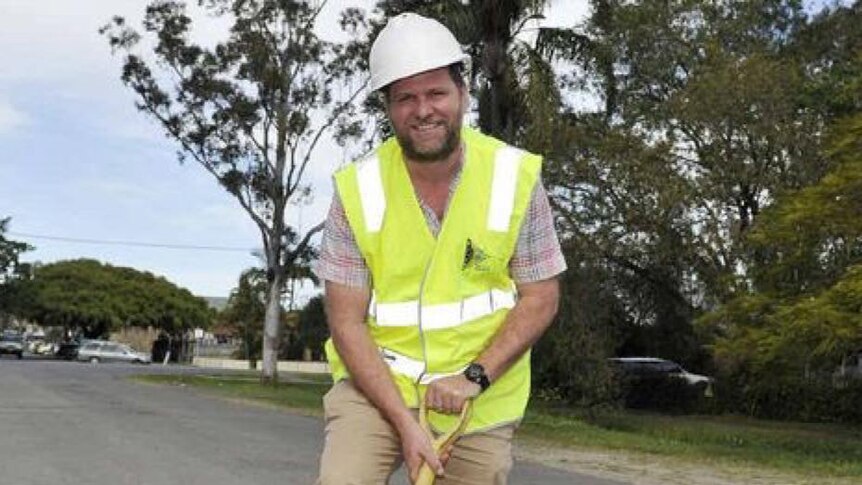 The Byron Shire mayor Simon Richardson wearing a high-vis vest and holding a shovel in a pothole on a sealed road