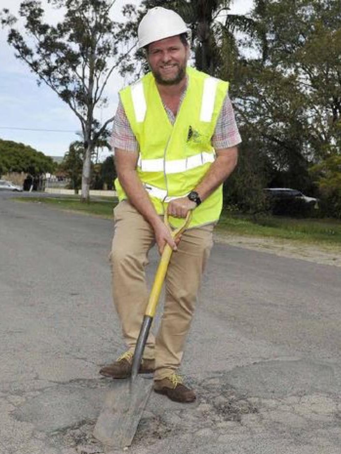 The Byron Shire mayor Simon Richardson wearing a high-vis vest and holding a shovel in a pothole on a sealed road