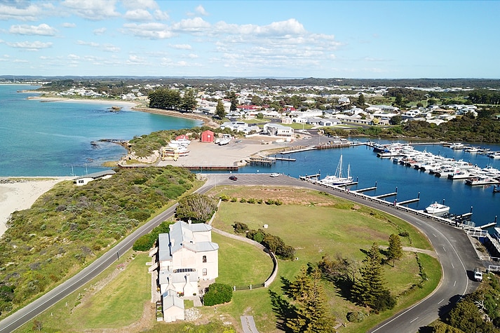 An aerial photo of a large estate on large well-kept lawns, overlooking a marina with docked boats on a sunny day.