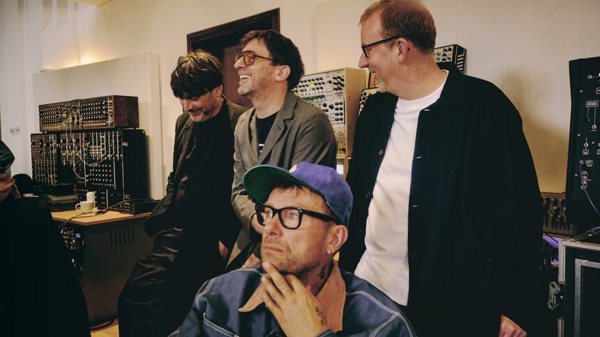 Blur listening in the studio, Damon Albarn sits in a chair contemplatively while the other three members share a laugh behind