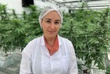 Emily Rigby wears a white coat and hair net in a greenhouse with cannabis plants in the background.