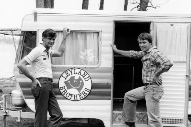 Black and white pic of two men in tee-shirt, jeans, other in checked shirt, jeans, lean on caravan with Leyland Brothers label.