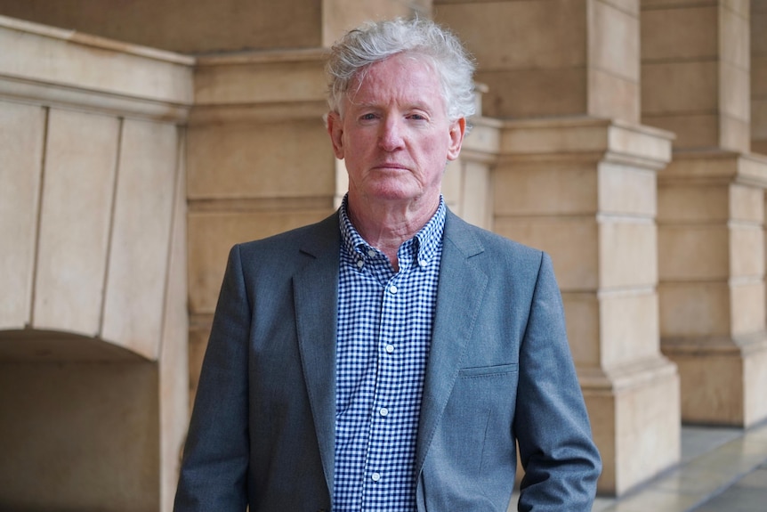 A man with grey hair wearing a suit jacket and blue checked shirt outside a stone building