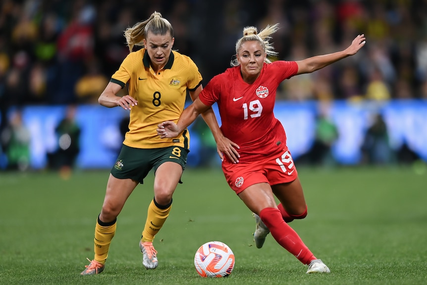 Two women soccer players, one wearing yellow and green and the other wearing red, battle for a ball during a game