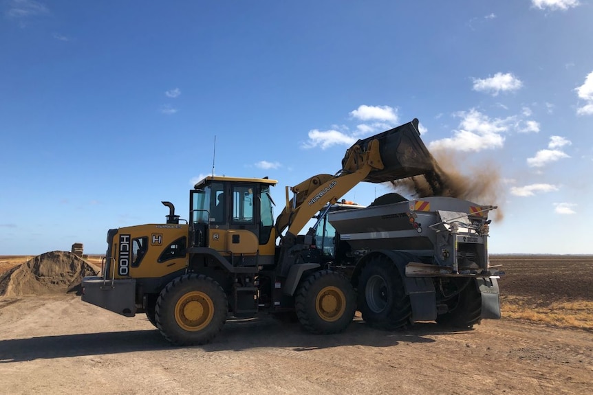 A yellow farm machine loads a brown, powdery material into a spreader.