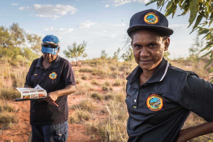 Two Indigenous people wearing ranger caps and shirts, stand in a scrubby grassland.