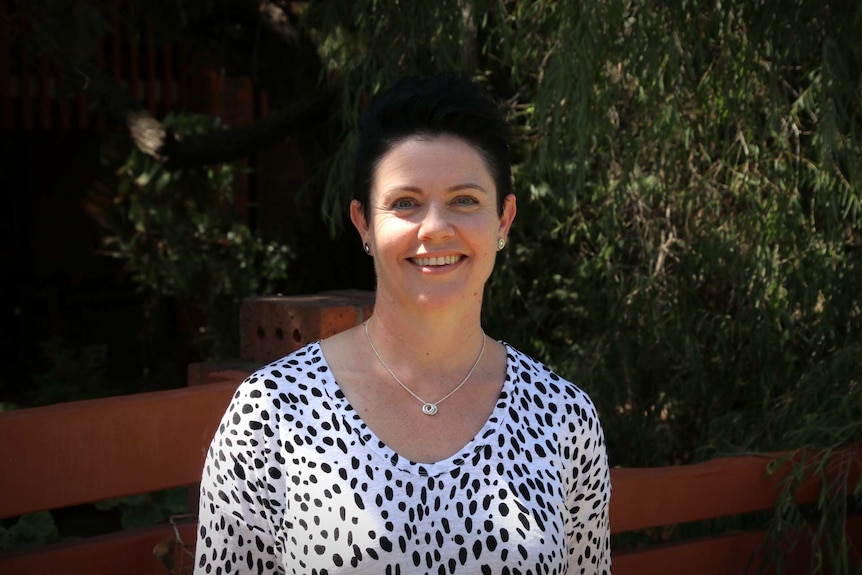 A woman with black hair and a necklace and black and white top stands in front of a brick wall and garden