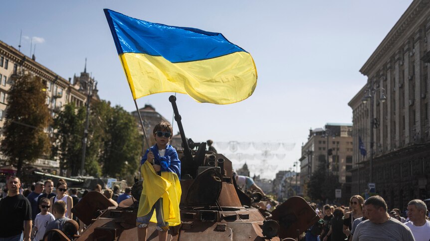 A young boy stands on top of a disused tank, waving a blue and yellow Ukrainian flag