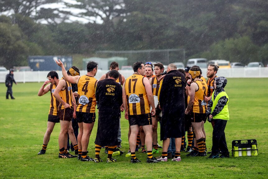 A huddle of male footballers in brown and gold jumpers stand on rainy football ground