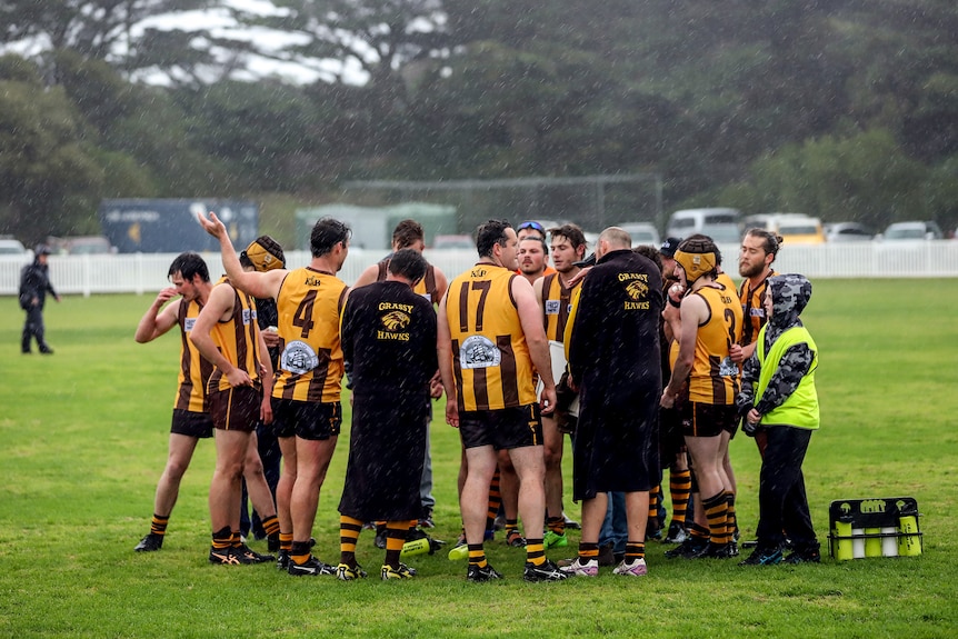 A huddle of male footballers in brown and gold jumpers stand on rainy football ground