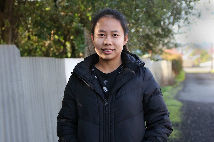 Young woman, Wah Ree Paw, stands, smiling, in front of a fence and tree.