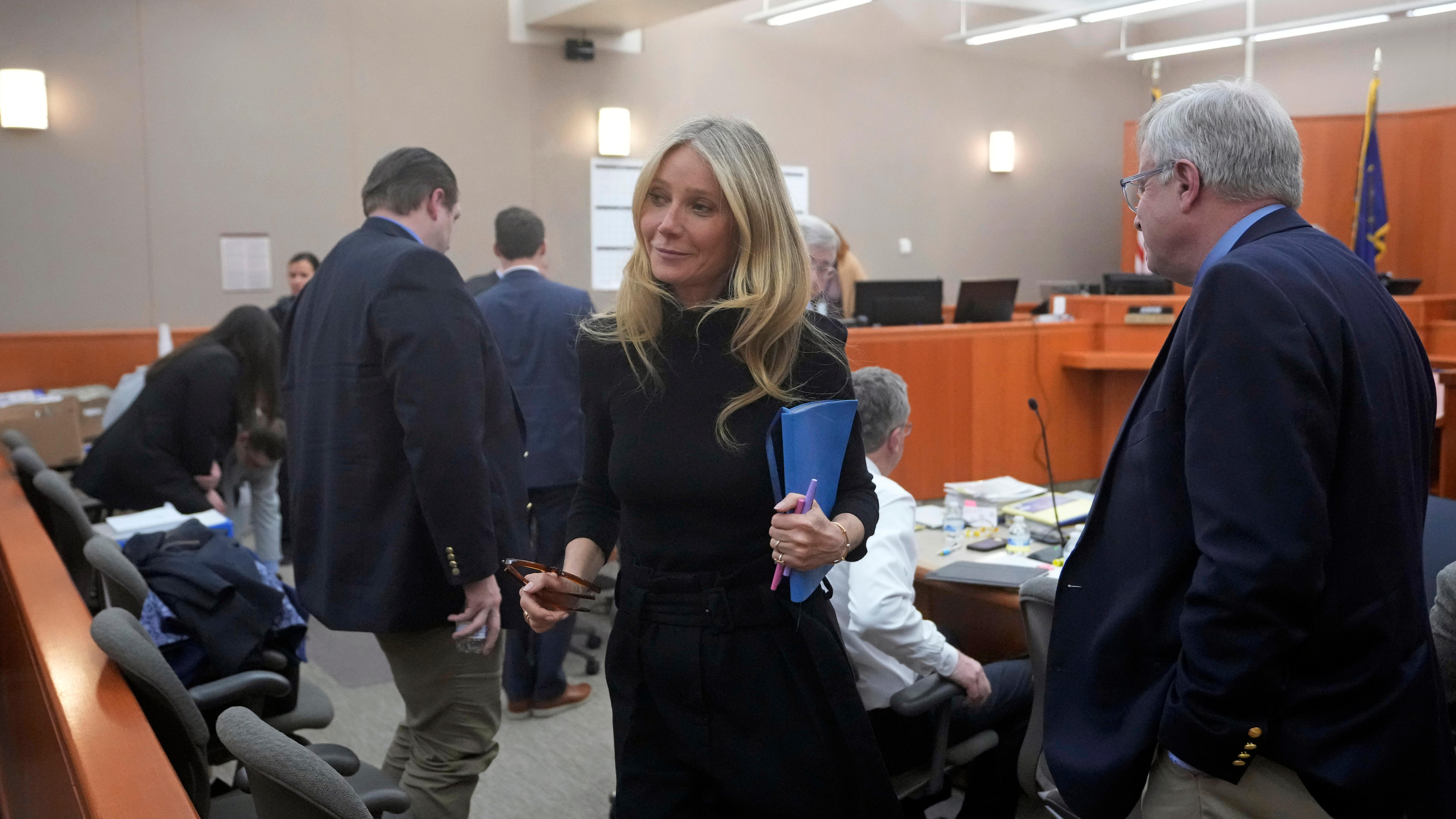A blonde woman in a courtroom, she holds her glasses and what looks like files and smiles at someone outside of the frame.