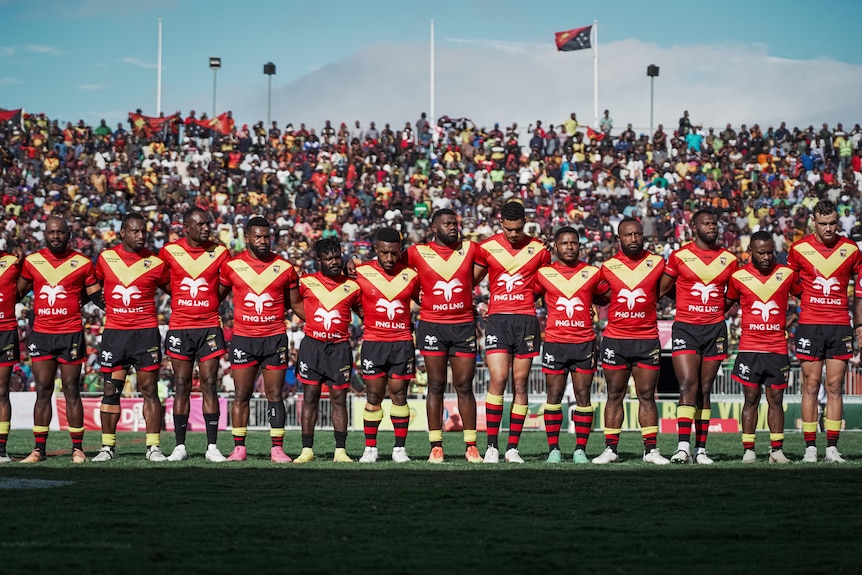 14 men are lining up on a field, behind them is a large crowd, they all wear red and yellow sport shirts and black shorts