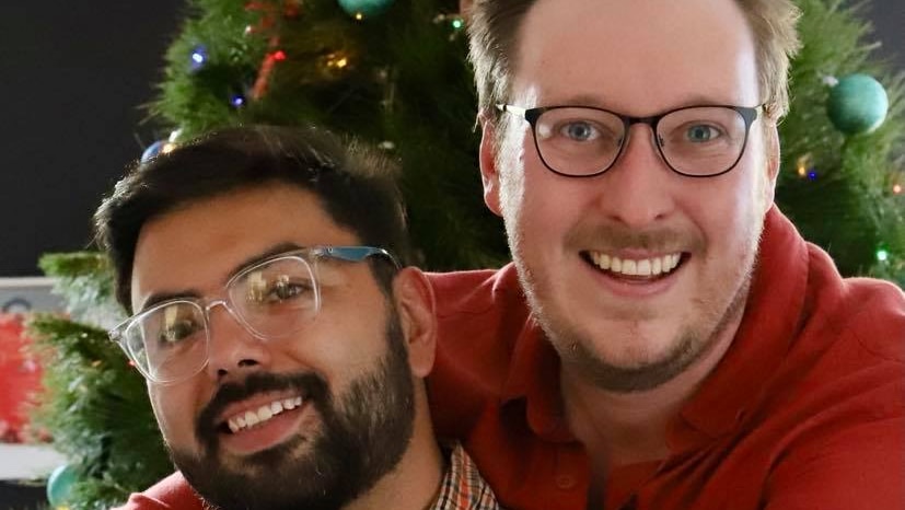 One man in a red shirt and glasses hugs another man in glasses in front of a decorative Christmas tree