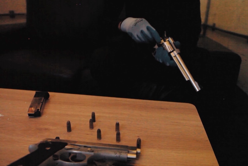 A gang member loads a revolver. In front of him on a table is a handgun and several bullets.