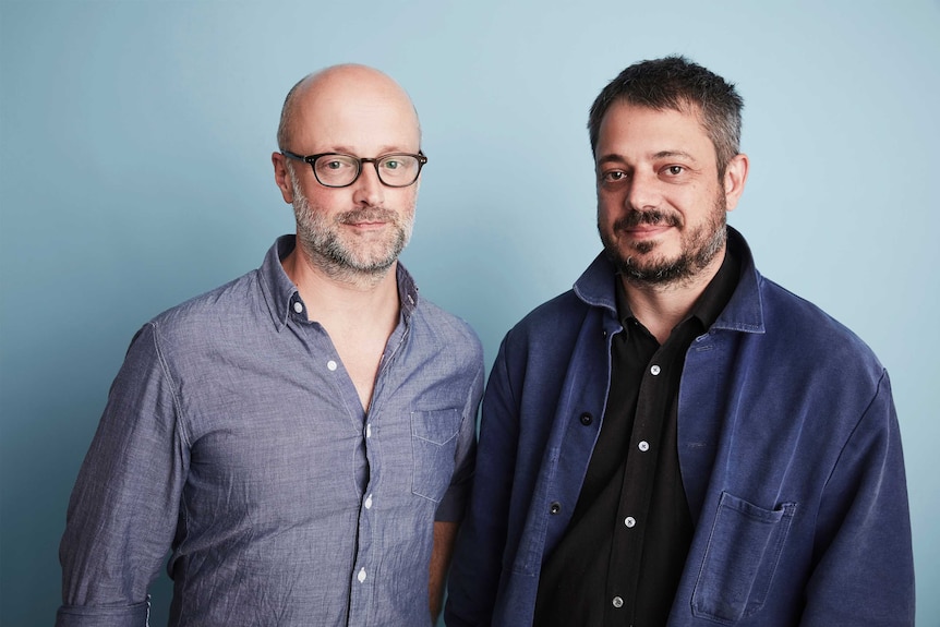 The two men pose in front of a light blue background.