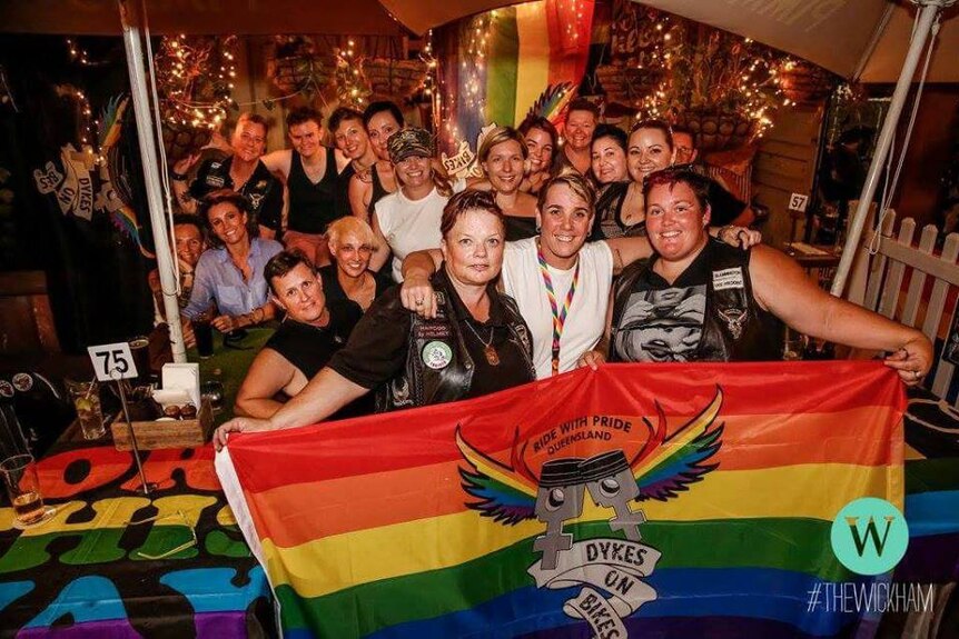 Women smile and hold up a rainbow flag with the Qld dykes on Bikes logo.