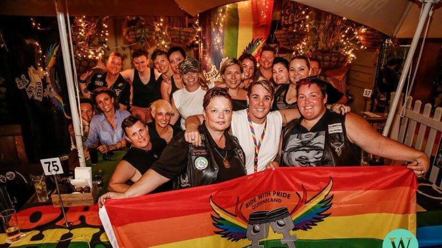 Women smile and hold up a rainbow flag with the Qld dykes on Bikes logo.