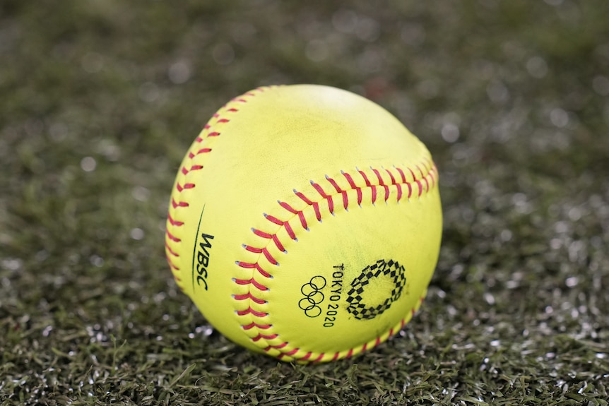 A yellow softball with a Tokyo 2020 logo sits on green grass
