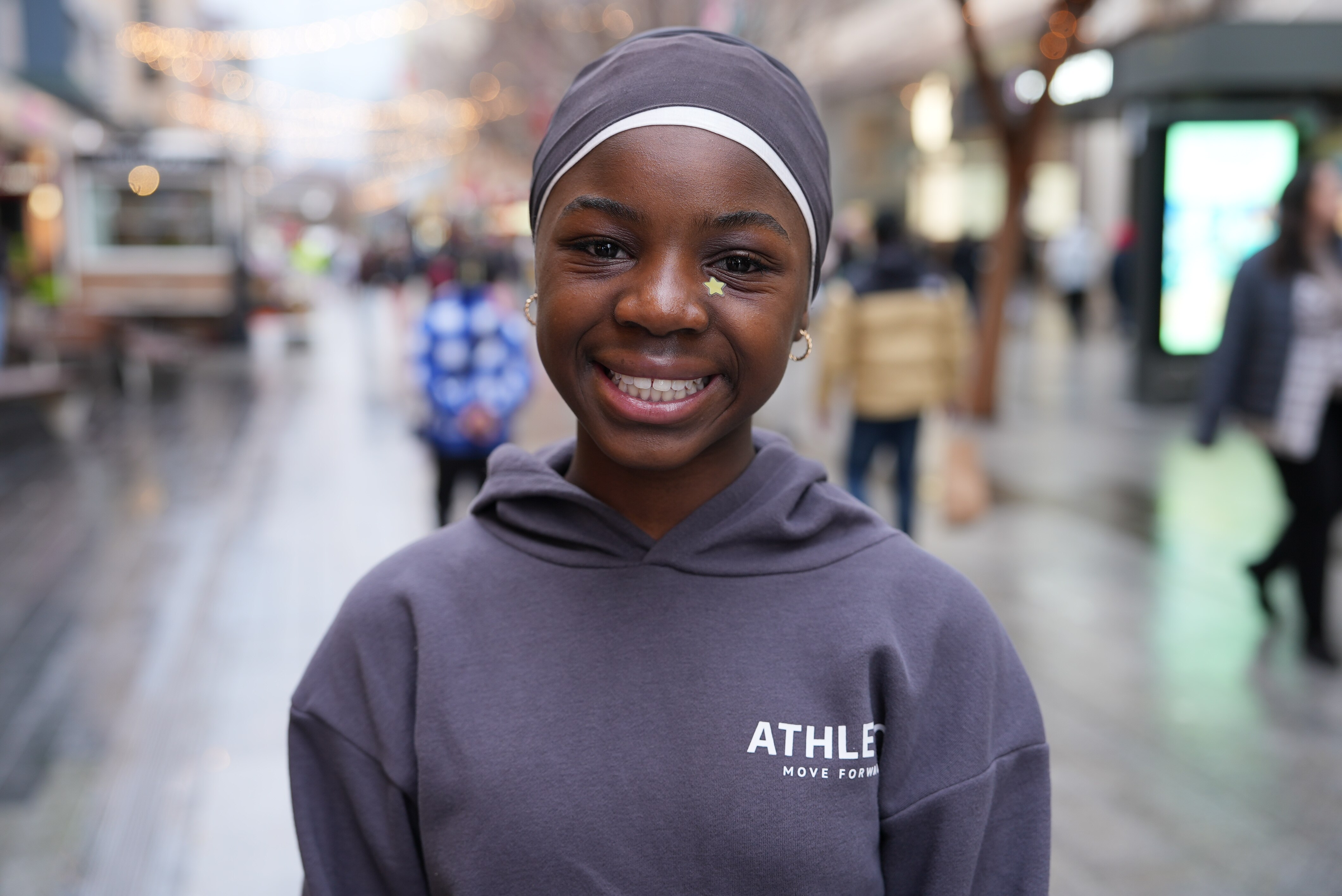 A smiling teenage girl with her hair covered by a scarf stands in an outdoor shopping mall where it has recently been raining.