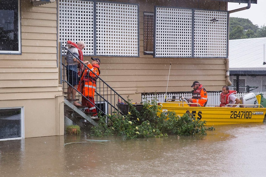 A resident stands on their front stairs of their flooded house being rescued by an SES crew.