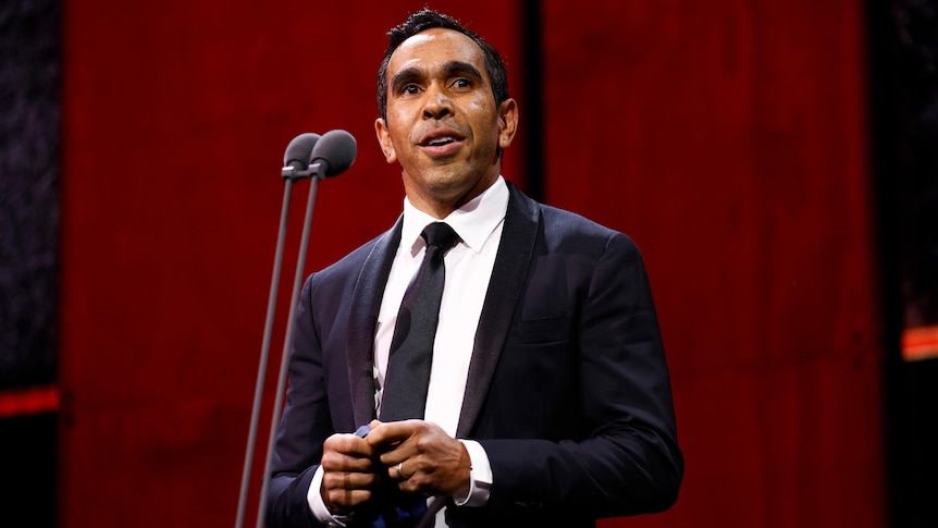 Former AFL player Eddie Betts stands in a suit on stage in front of a microphone during an awards ceremony.