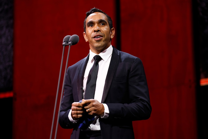 Former AFL player Eddie Betts stands in a suit on stage in front of a microphone during an awards ceremony.