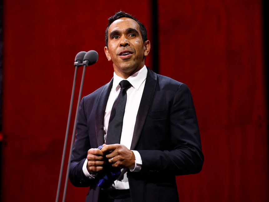 Former AFL player Eddie Betts stands in a suit on stage in front of a microphone during an awards ceremony.