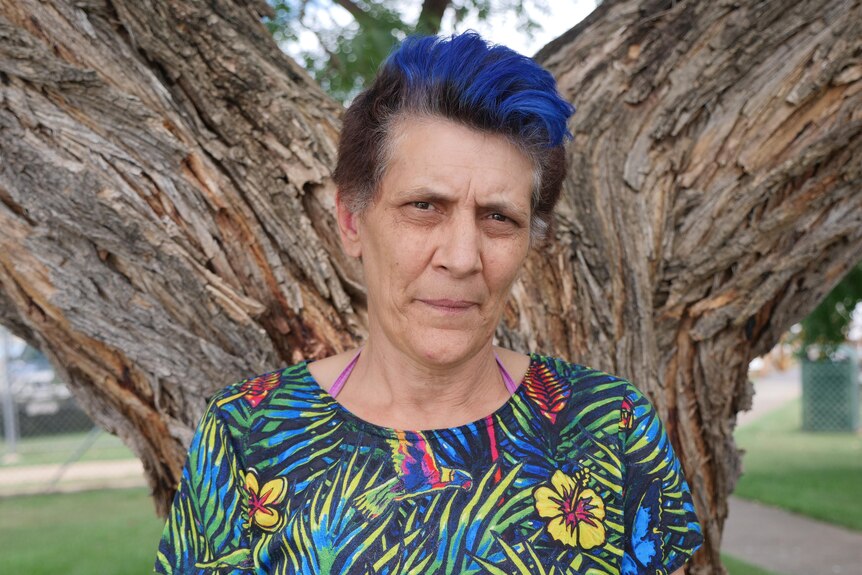 A middle-aged woman with short, dyed hair, wearing a colourful top, stands in front of a tree.