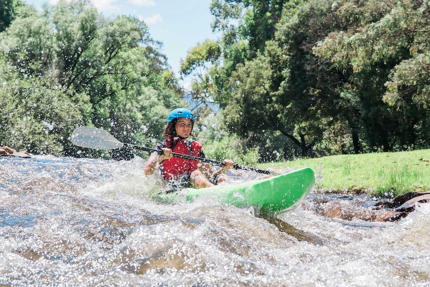 A woman in a blue helmet and red flotation vest kayaks down a river in white water
