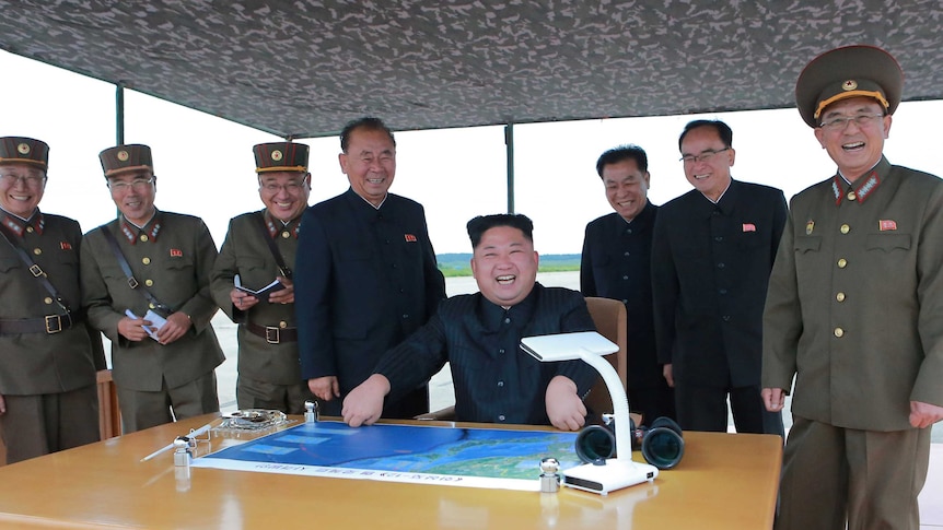 Kim Jong-un sits over a map and laughs and men standing around him laugh too.