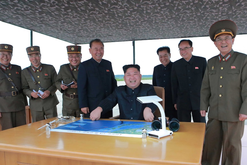 Kim Jong-un sits over a map and laughs and men standing around him laugh too.
