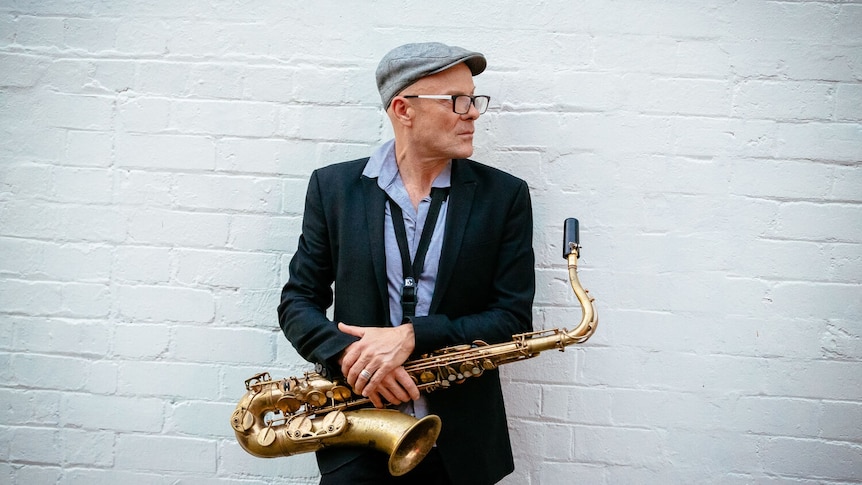 Jason Bruer stands in front of a white brick wall with his bronze tenor sax