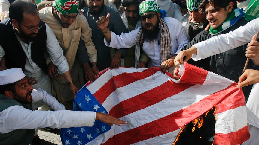 A group of bearded men in traditional Pakistani clothing burn a US flag.