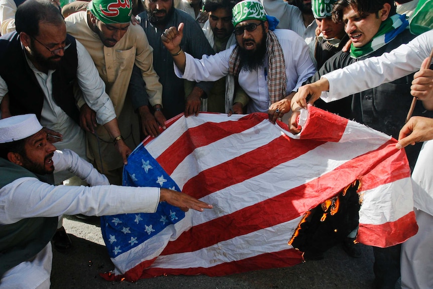 A group of bearded men in traditional Pakistani clothing burn a US flag.