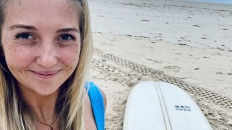 A young women with long blonde hair is smiling while standing on a beach with a surf board on the sand behind her.