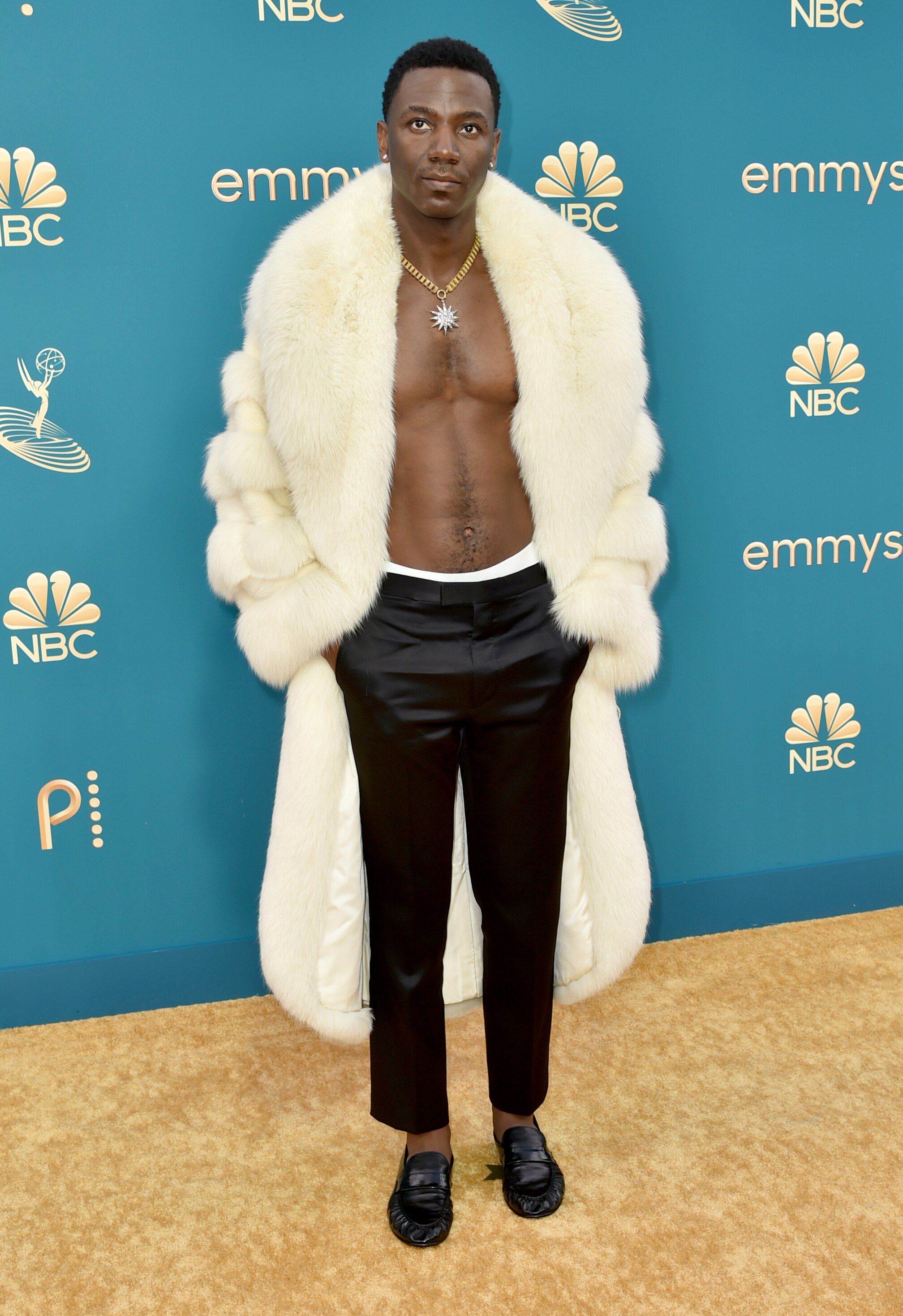 Jerrod Carmichael wearing an open white fur coat, a starbust necklace and silky black trousers. 