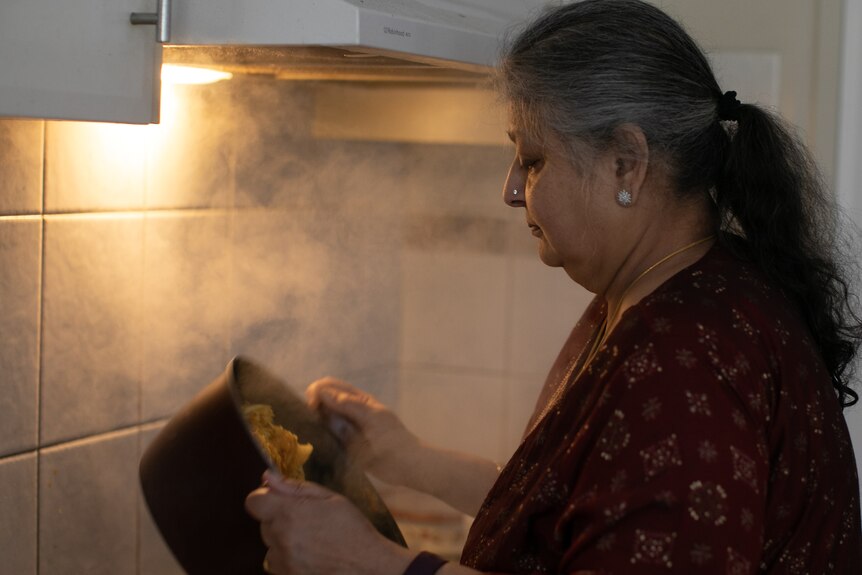 An older woman with salt and pepper hair tied back, nose pin, earings, looks at a steaming pot she holds, tiled background.